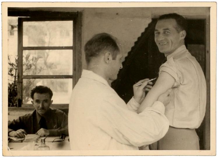 Black-and-white photograph of a man holding a scalpel to the upper arm of another man who is smiling for the camera