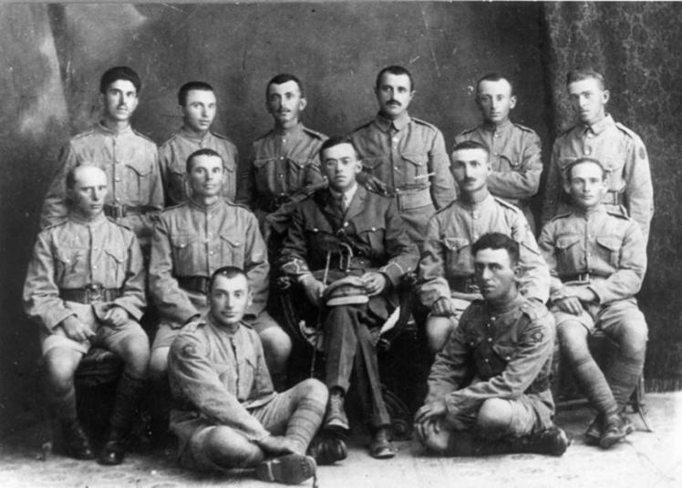 Gruppenbild von zwölf uniformierten Männern, wobei die Person in der Mitte mit Brille, Stock und Mütze auf dem Schoss hervorsticht.