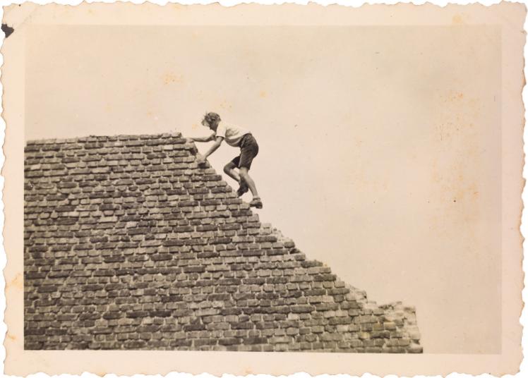 Black-and-white photograph of a young man wearing shorts and a white shirt, recklessly climbing a firewall