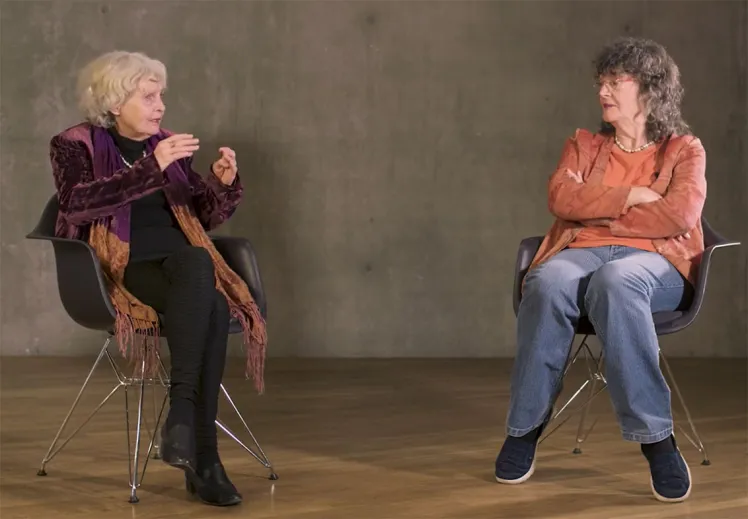 Two elderly ladies are sitting on chairs in front of a concrete wall, chatting.