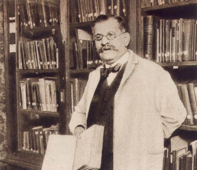 Schwarz-weiß-Foto: Magnus Hirschfeld mit Brille, Schnurbart und weißem Kittel. Er steht vor einem Buchregal und schaut freundlich in die Kamera. 