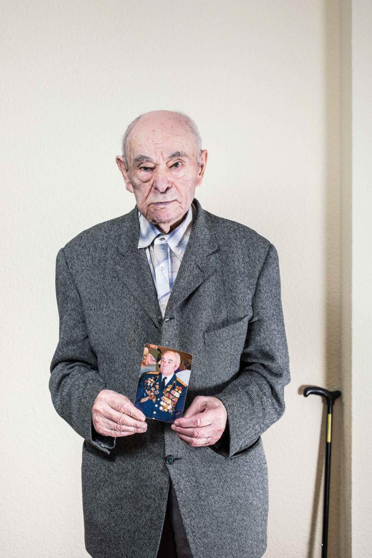 elderly man in grey suit holding a photo of himself covered in medals and decorations
