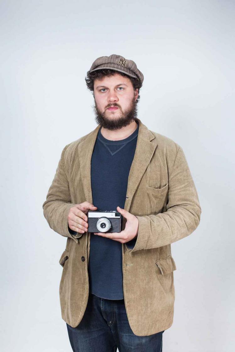 A young man with a cap and beard in a brown corduroy jacket is holding an analogue camera in his hands.