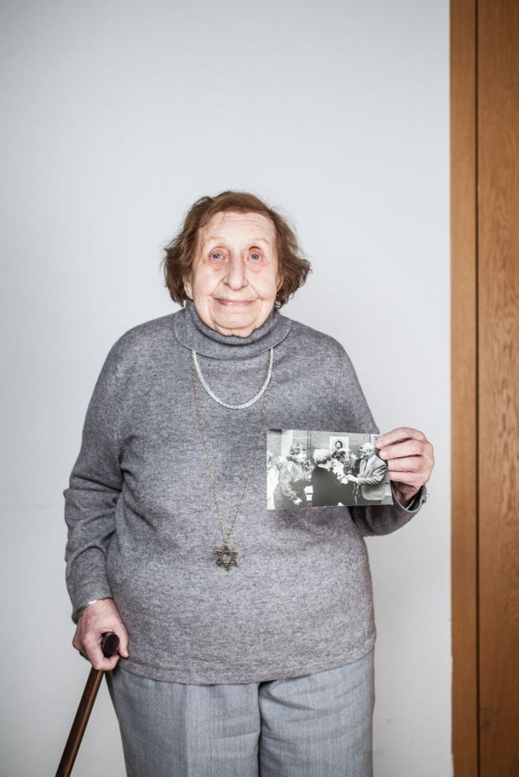 Eine Frau stützt sich auf einen Gehstock und hält zwei Fotos in der Hand.