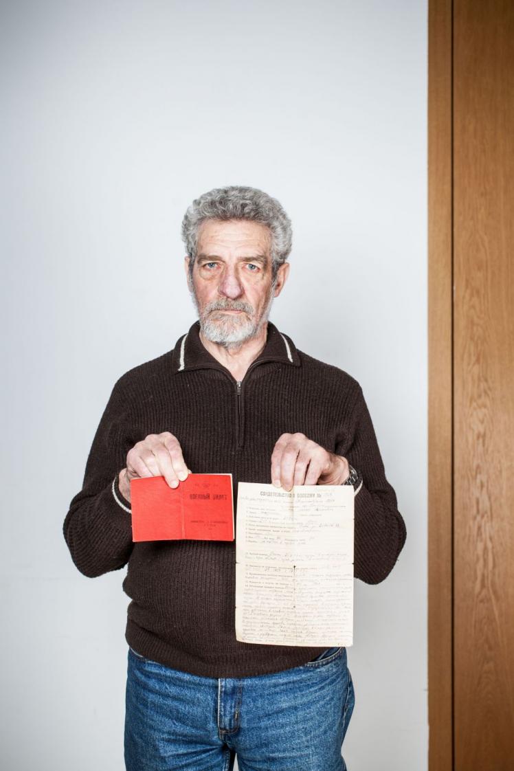 A bearded gray-haired man holds up two documents