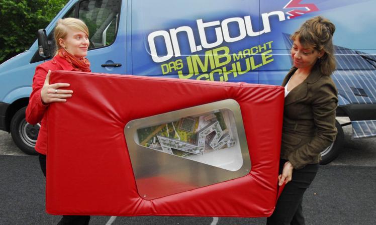 Zwei Mitarbeiterinnen von on.tour tragen einen roten Sitzwürfel, in dem ein Modell des Libeskind-Baus vom Jüdischen Museumzu sehen ist.