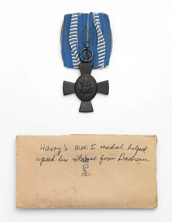 Metallenes Kreuz mit der Aufschrift 7.1.1916 am blauen Band, darunter ein Zettel mit handschriflicher Notiz.
