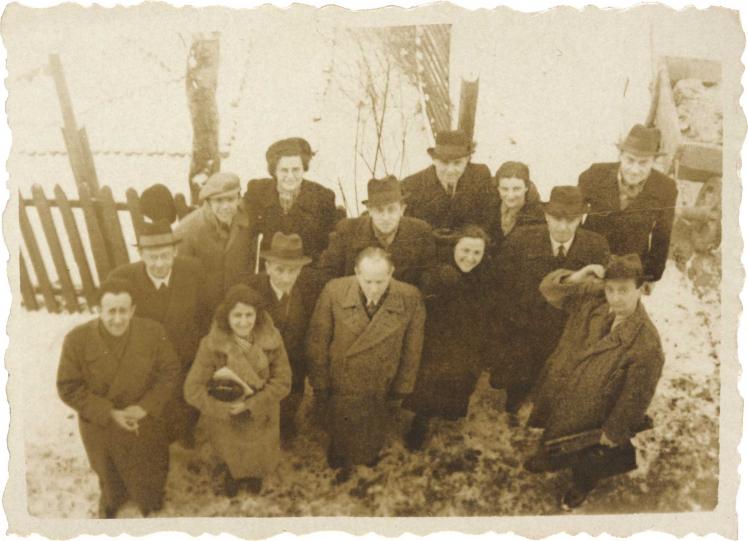 Ausgewaschenes Schwarz-Weiß-Foto einer Gruppe von 14 Männern und Frauen in Mänteln. Sie stehen auf einer schneebedeckten Fläche vor einem Zaun und wurden von oben fotografiert. Fast jeder schaut in die Kamera, einige lächeln.