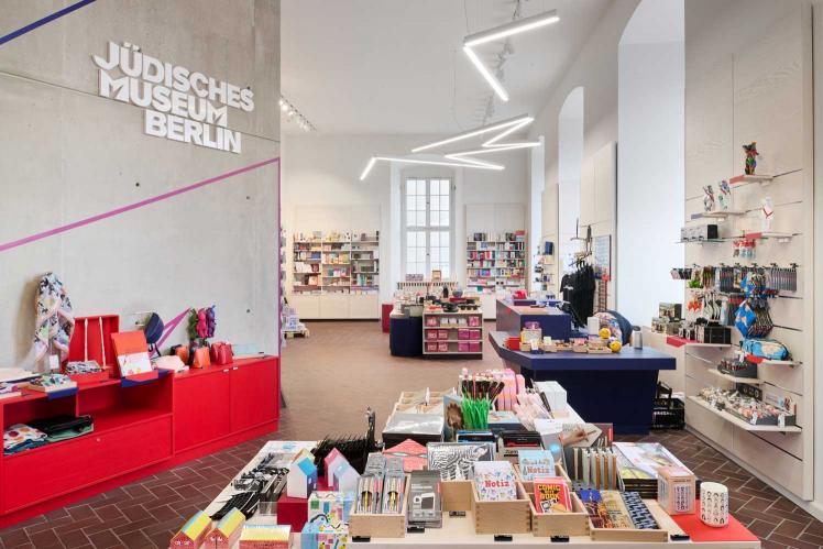  Espace boutique avec des meubles aux couleurs vives et un assortiment coloré, le logo blanc avec l'inscription Jewish Museum Berlin peut être vu sur un mur en béton et une lampe au néon au plafond qui zigzague dans la pièce.