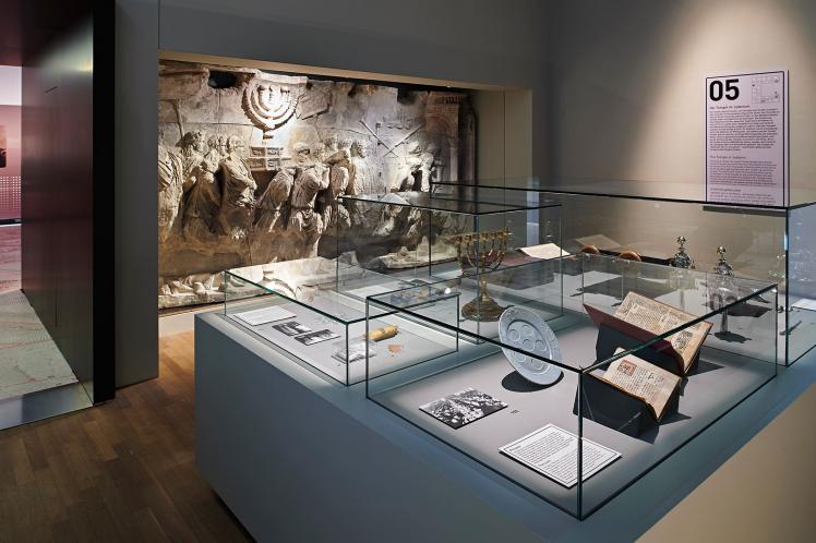 A look into an exhibition room. On the rear wall, a plaster cast of the Arch of Titus can be seen. In the foreground stand a glass case with a menorah, Torah rimonim, and open books