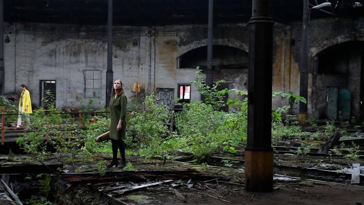 A woman stands in the middle of an old decaying building and looks up, she is surrounded by plants and remains of the building