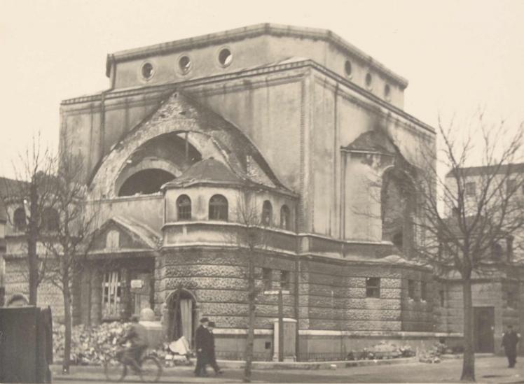 View of the corner of the burnt-out synagogue, in front of the building bare trees and passers-by are recognizable.