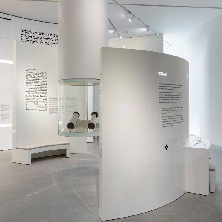 Round showcase hanging from the ceiling with a Torah scroll inside. To the right of the showcase is a curved wall on which an introductory text about the Torah is printed. The Ten Commandments are printed in Hebrew on a curved wall to its left.