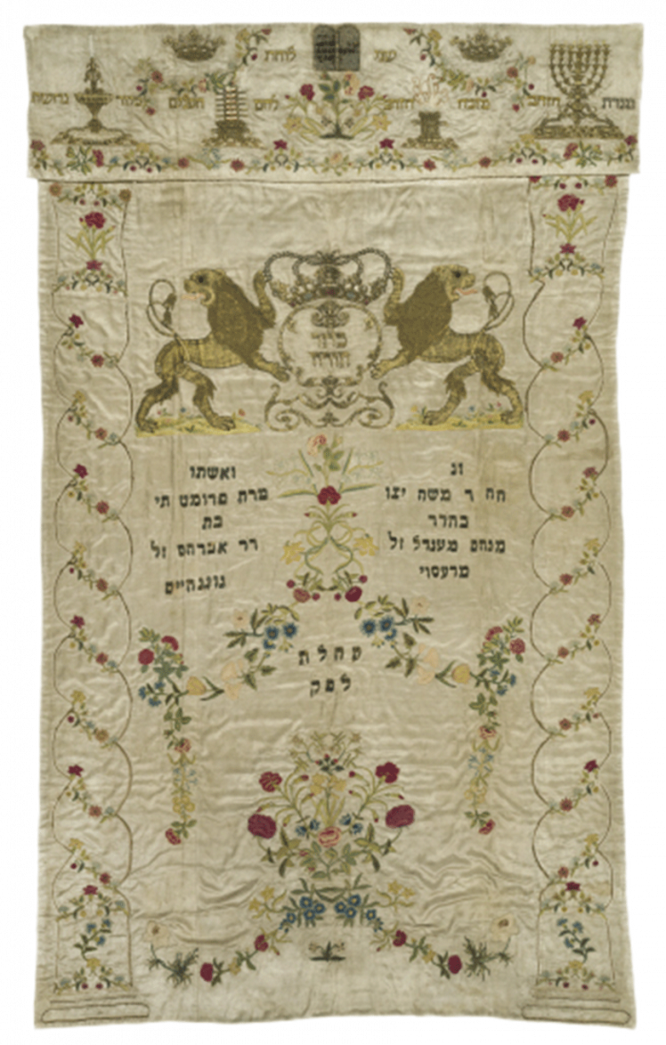 Embroidered white Torah curtain with numerous decorations and two lions and a crown over the Hebrew dedication inscription.