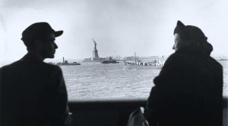 A man and a woman looking at each other, in semi-profile, on a ship, with a clear view over the water to the Statue of Liberty 
