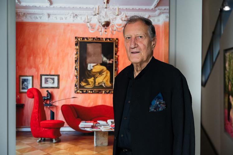 An elder man with a pocket square in a jacket stands in front of a large format photo of a salon where a painting of a woman hangs on the wall, where the head would be, a piece of canvas is cut out