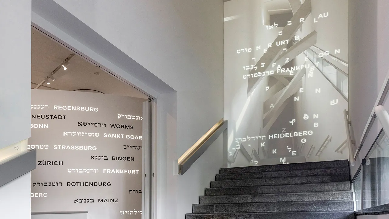 Eine Treppe in der Dauerausstellung. An den Wänden stehen Ortsnamen in hebräischer und lateinischer Schrift.
