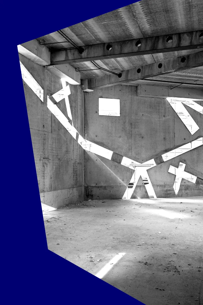 Schwarz-wei&szlig;-Foto eines noch im Bau befindlichen, leeren Raums, in dessen Betonw&auml;nden sehr schr&auml;ge Fenster sind, durch die Licht einf&auml;llt.