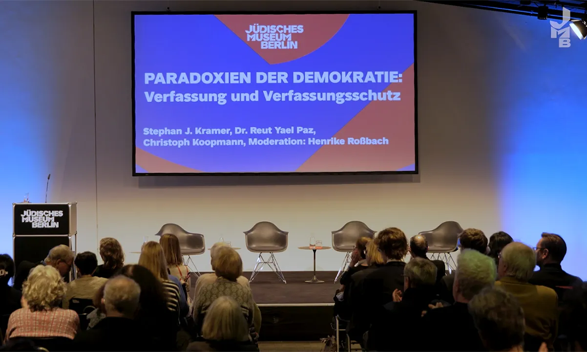 The audience, visible from behind, is looking at an empty stage with four chairs and a table. Behind them hangs a screen on which a presentation is projected. On the left is a lectern bearing the logo of the Jewish Museum Berlin.