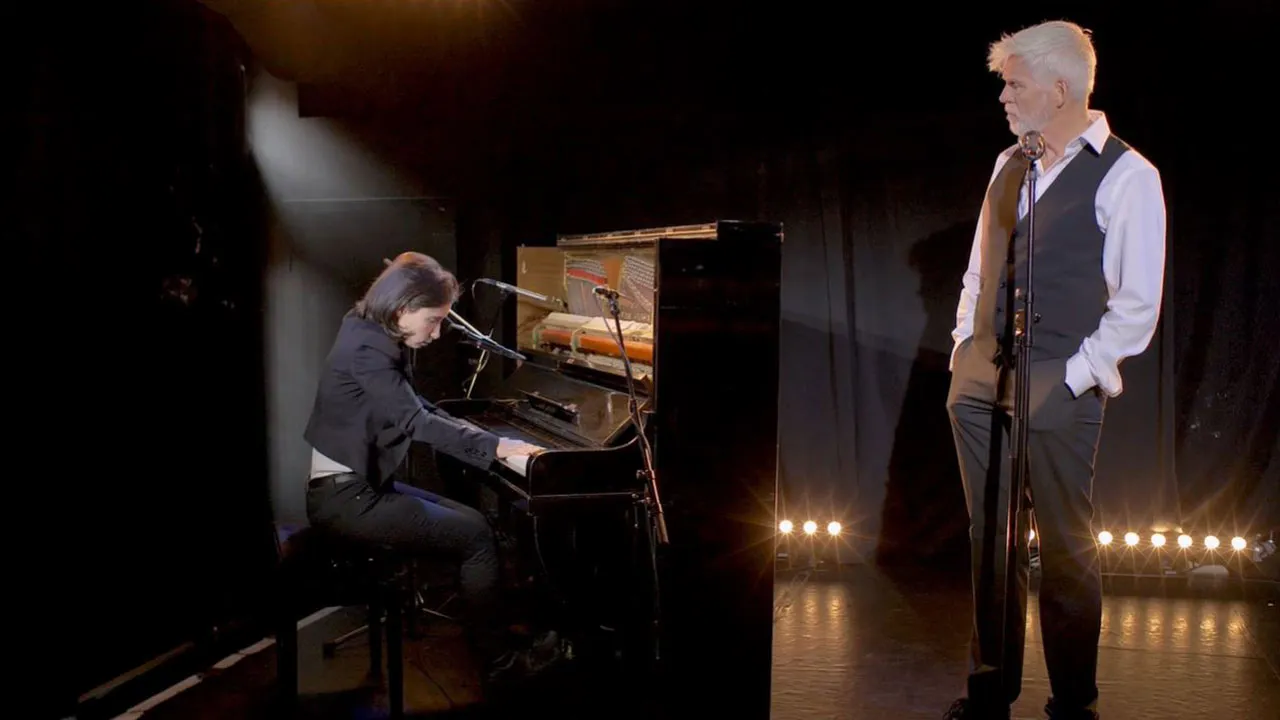 On a stage with black curtains in the background: a young woman plays the piano, a slightly older man stands next to her at the microphone and looks at her.