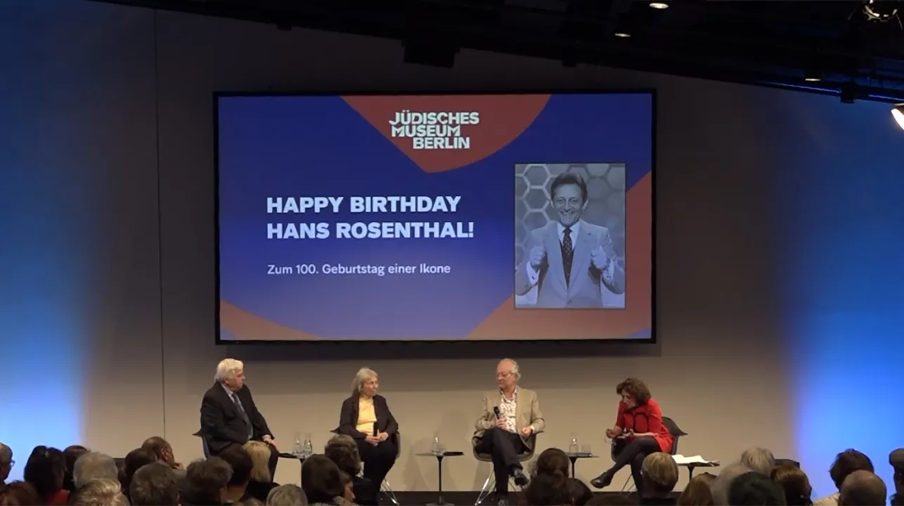 Two men and two women are sitting on a podium in front of an audience. Behind them is a screen on which a film is projected showing a photograph on the right and the words “Happy Birthday, Hans Rosenthal” on the left. 